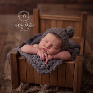May include: A newborn baby sleeping in a wooden bed with a gray knitted blanket and a gray knitted hat with a pom-pom.