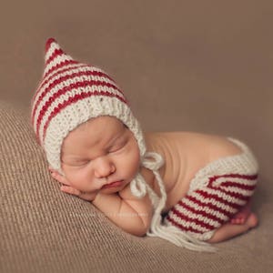 May include: A newborn baby wearing a knitted hat and shorts set. The hat is white with red stripes and a pointed top. The shorts are white with red stripes. The baby is lying on a brown surface.