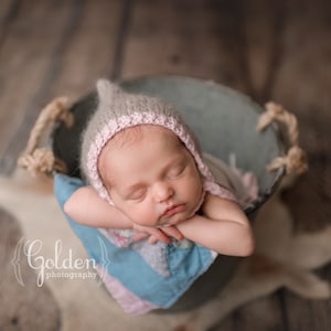 May include: A newborn baby wearing a grey and pink knitted hat sleeps in a metal bucket with a blue and pink blanket. The bucket has rope handles.