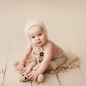 May include: A baby wearing a cream knitted hat and matching outfit sits on a cream textured surface, holding a cream knitted teddy bear.