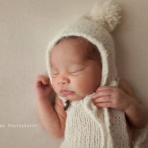 Könnte beinhalten: Ein schlafendes Baby, das eine weiße Strickmütze mit Bommel und ein passendes Strickoutfit trägt.