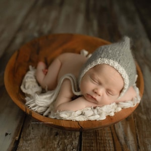 May include: A newborn baby wearing a grey knitted hat with a white trim sleeps peacefully in a wooden bowl. The baby is wrapped in a white blanket and is lying on its side.