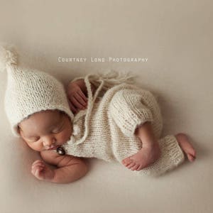 May include: A newborn baby wearing a white knitted hat and matching knitted outfit with a brown button. The baby is sleeping on a light brown background. "COURTNEY LONG PHOTOGRAPHY" is written in white text at the top of the image.
