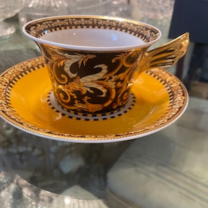 May include: A decorative teacup and saucer set. The cup features a black and gold design with a winged handle. The saucer is yellow with a matching gold border. The set is likely made of porcelain.