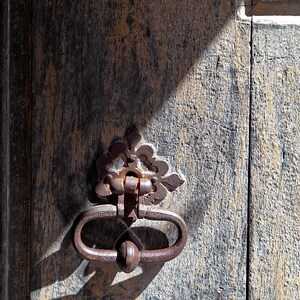 May include: A close-up of an ornate, rusty metal door knocker with a decorative design. The knocker is attached to a weathered wooden door.