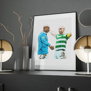 Puede incluir: Una impresión enmarcada que representa a dos futbolistas, uno con una camiseta azul claro y el otro con rayas verdes y blancas. La obra de arte se exhibe sobre una superficie oscura con lámparas y jarrones decorativos.