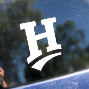 May include: A white vinyl decal of a stylized letter H with a white swoosh underneath. The decal is on a blue car window.