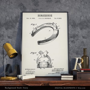 May include: A black and white patent drawing of a horseshoe design. The drawing shows the horseshoe in detail, with labels and numbers indicating different parts of the horseshoe. The patent information is included at the top of the drawing, including the date, inventor's name, and patent number. The drawing is framed and displayed on a wooden surface with a lamp and other decorative items.