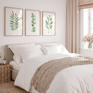 May include: A white bed with a beige knitted throw blanket and white pillows. Three framed watercolor paintings of green leaves hang on the wall behind the bed.