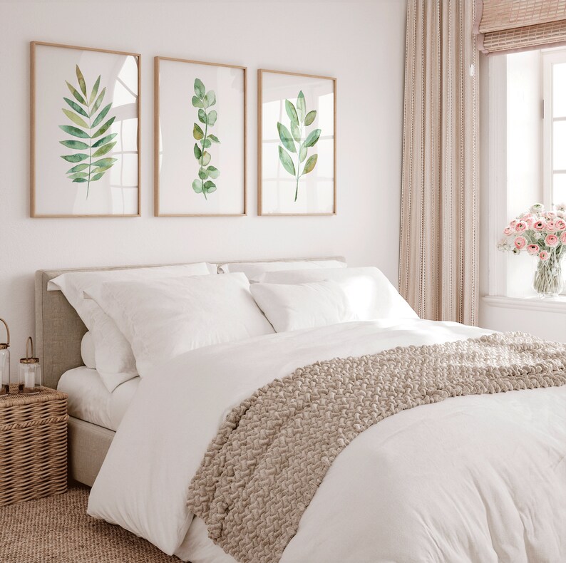 May include: A white bed with a beige knitted throw blanket and white pillows. Three framed watercolor paintings of green leaves hang on the wall behind the bed.