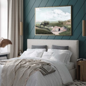 May include: A painting of a landscape with a large stone blocking the entrance to a cave. The painting is framed in gold and hangs on a teal wall above a bed with white bedding and a beige throw blanket.