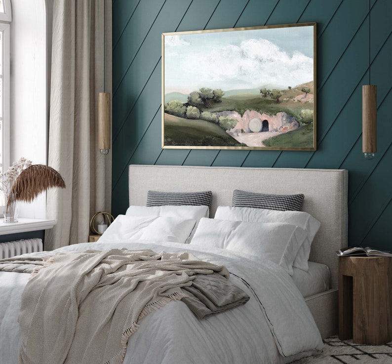 May include: A painting of a landscape with a large stone blocking the entrance to a cave. The painting is framed in gold and hangs on a teal wall above a bed with white bedding and a beige throw blanket.