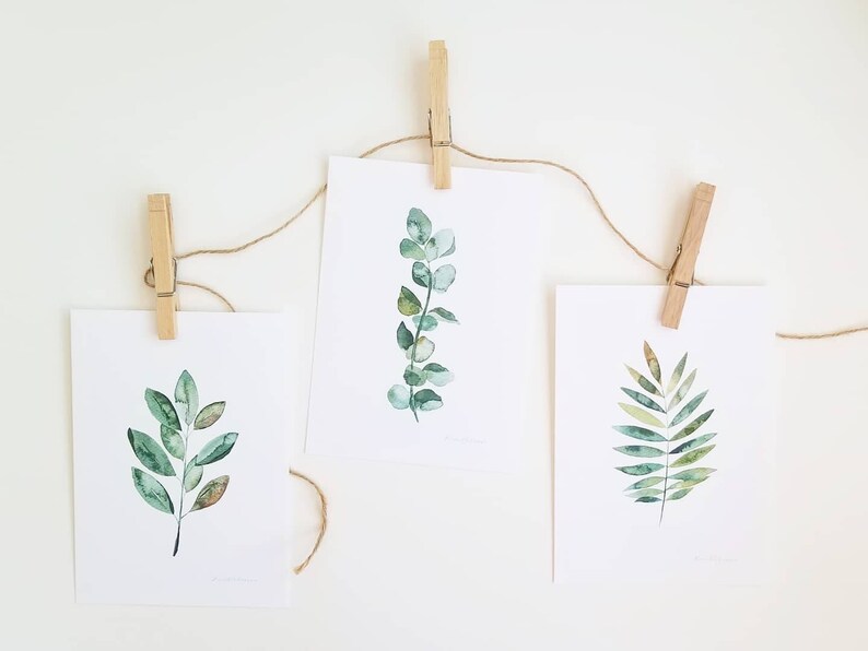 May include: Three watercolor paintings of green leaves are displayed on a white wall using twine and wooden clothespins. The leaves are painted in shades of green and brown.