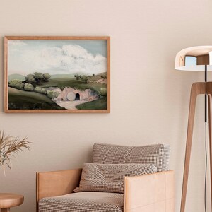 May include: A framed painting of a landscape with a large stone tomb in the foreground. The painting is in a light wood frame and is hanging on a light beige wall. A brown leather armchair with a checkered cushion sits in front of the painting.