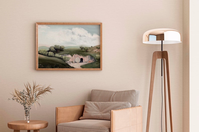 May include: A framed painting of a landscape with a large stone tomb in the foreground. The painting is in a light wood frame and is hanging on a light beige wall. A brown leather armchair with a checkered cushion sits in front of the painting.