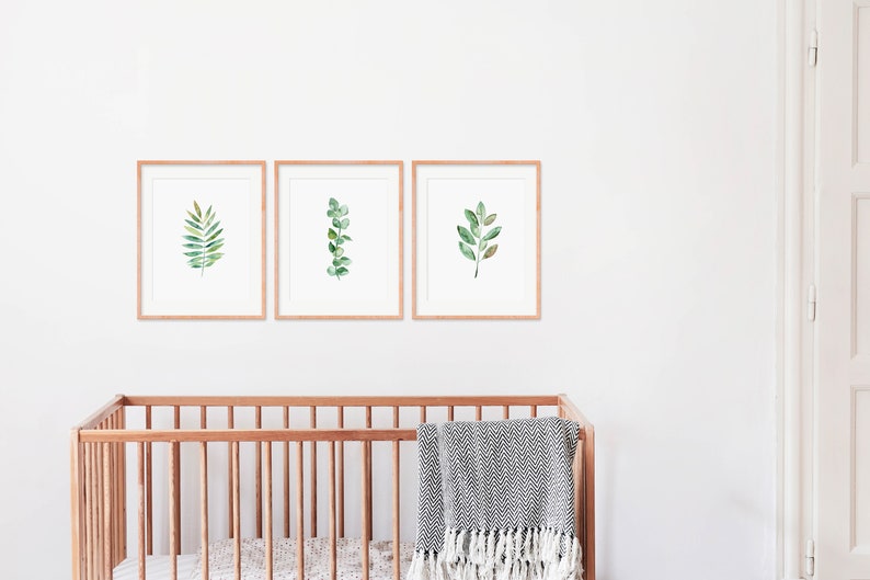 May include: Three framed watercolor prints of green leaves on a white wall above a wooden crib with a gray and white blanket.