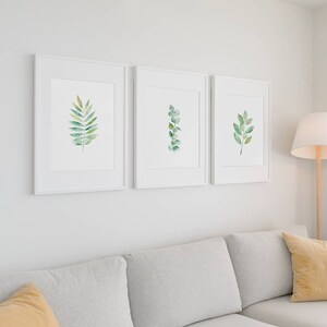 May include: Three white framed prints of watercolor illustrations of green leaves on a white wall above a light gray couch with two yellow throw pillows.