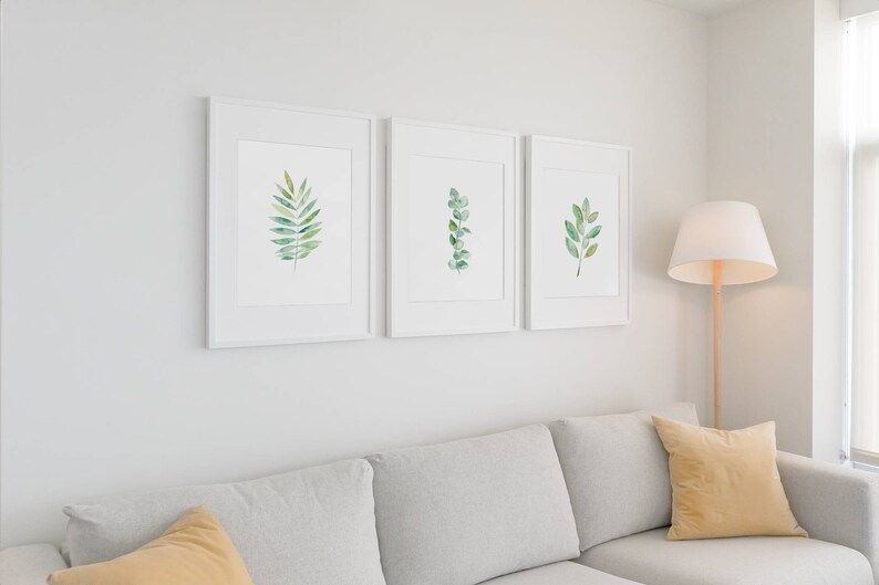 May include: Three white framed prints of watercolor illustrations of green leaves on a white wall above a light gray couch with two yellow throw pillows.
