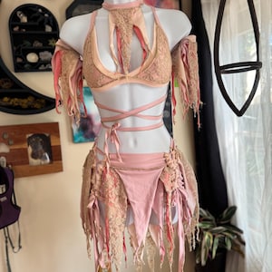 Puede incluir: Conjunto de lencería de dos piezas en tonos rosa y melocotón, exhibido en un maniquí. El conjunto presenta un top halter, puños en los brazos y una falda con detalles de encaje y cintas. El diseño tiene una estética bohemia o de hadas.