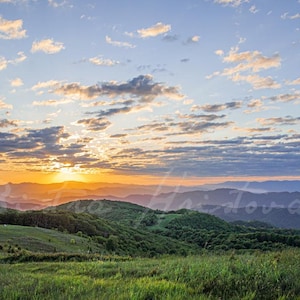 May include: A scenic landscape photograph of a mountain range at sunset. The sky is filled with clouds, and the sun is setting behind the mountains, casting a warm glow. The foreground features green grass and rolling hills.
