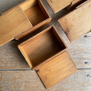 May include: Several small, open wooden boxes with a natural finish are arranged on a wooden surface. The boxes are rectangular with hinged lids, and some are open, revealing the interior. The wood grain is visible.