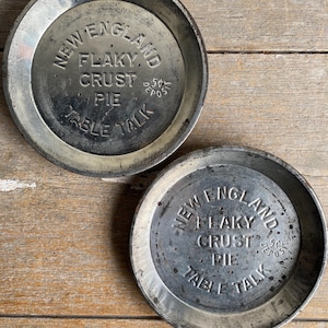May include: Two antique metal pie plates, each with the embossed text "NEW ENGLAND FLAKY CRUST PIE TABLE TALK". The plates are round with a raised rim and show signs of age and use, typical of vintage items.