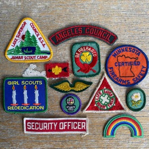 May include: A collection of twelve embroidered fabric patches featuring various shapes and designs, including text such as Lake Superior Council B.S.A., Angeles Council, Minnesota Certified Firearms Safety, Girl Scouts Rededication, and Security Officer.