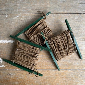 May include: Three wooden rope spools with green frames and tightly wound brown rope. The spools are arranged in a triangular pattern on a weathered wooden surface, showcasing a rustic aesthetic. The rope is secured with knots.
