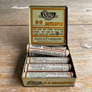 May include: A vintage tin box of Ansco Cyko O-H Developer for photographic film and paper. The box contains six individual tubes of developer, each labelled with instructions for use. The box is labelled with the brand name "Ansco Company" and the location "Binghamton, N.Y. U.S.A."