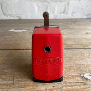 May include: A vintage red pencil sharpener with a black base and a metal crank. The sharpener has a round hole for pencils and the text "BOSTON, Mass." and "TO REMOVE" on the front.