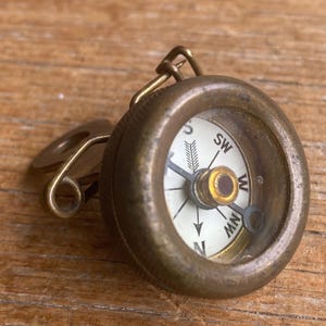 May include: A small, round brass compass featuring a white dial with black directional markings including N, S, SW, W, and NW. The compass has a clear glass cover and a metal pin attachment on the side, designed for securing the device to fabric.