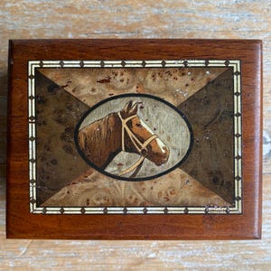 May include: A rectangular wooden box featuring a central oval inlay of a brown horse head wearing a bridle. The lid displays a geometric pattern with dark and light wood veneers, accented by a thin white border with a repeating diamond design.