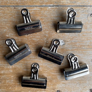 May include: Six silver-toned metal binder clips, each featuring a rectangular base and a hinged handle with a circular opening. The clips are embossed with the text "BOSTON CLIP NO. 4" and "HUNT MFG. CO." on the front surface of the metal body.