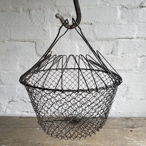 May include: A dark grey metal wire basket with a conical shape and a fine mesh construction. The basket features a wide circular rim and a tapered base, suspended from a central metal hook by three thin wire supports that meet at the top.
