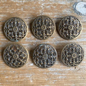 May include: A set of six circular brass buttons featuring an intricate openwork design. Each button displays a central floral motif surrounded by a square frame and delicate filigree scrollwork, finished with a dark, aged metallic patina.