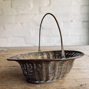 May include: An antique metal basket with a woven pattern and a curved handle. The basket has a dark, aged look, indicating it's vintage. The handle curves elegantly above the oval body, enhancing its classic charm. The basket rests on a wooden surface.