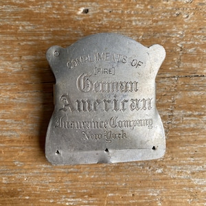 May include: A silver-toned metal promotional item with an arched top and scalloped sides. The text reads: COMPLIMENTS OF [FIRE] German American Insurance Company New York. The object features small triangular cutouts along the bottom edge.
