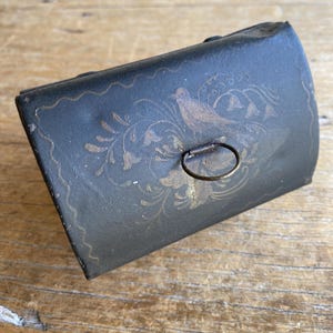 May include: A small, antique black metal box with a hinged lid and a metal ring closure. The box is decorated with a gold-coloured bird and floral design. The edges of the box have a decorative wavy pattern.