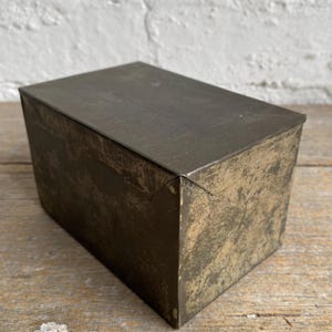 May include: A rectangular metal box with a weathered, dark grey and bronze-toned finish. The container features a flat, hinged lid that covers the top, with visible signs of oxidation and surface wear across its exterior panels and edges.