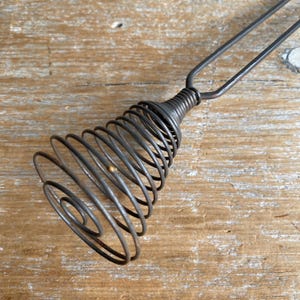 May include: A vintage metal whisk with a spiral design. The whisk has a series of concentric coils that narrow towards the handle. The handle is made of two parallel rods that join the coiled section. The whisk is made of a dark, possibly iron, metal.