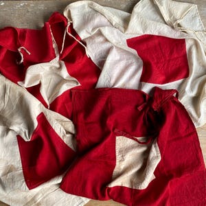 May include: A collection of fabric flags featuring a bold geometric design with alternating large red and off-white square panels. The textile pieces are made of cotton, showing visible stitching along the seams and integrated fabric ties.