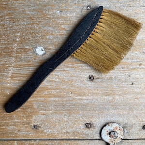 May include: A black-handled brush with a rectangular head of light brown bristles. The brush is angled across a weathered wooden surface, showcasing its design and texture. The handle appears to be made of leather or a similar material.