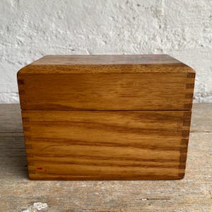 May include: A rectangular wooden box with a natural light brown grain and visible finger joints at the corners. The box features a flat lid and a smooth finish, showcasing the organic texture of the wood with a small red mark on the lower front.