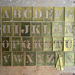 May include: A complete set of rectangular brass stencils featuring the uppercase English alphabet from A to Z and an ampersand symbol. Each individual metal plate contains a single cutout letter or character with a distinct serif typeface design.