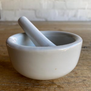 May include: A cream-coloured ceramic mortar and pestle. The mortar is a rounded bowl, and the pestle is a cylindrical rod. This set is for grinding and mixing ingredients, often used in cooking and traditional medicine.