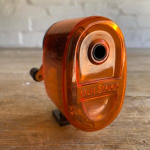 May include: Orange plastic pencil sharpener with a black crank and the word "BULLDOG" printed on the side.