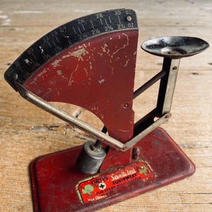 May include: A vintage red and black scale with a curved measurement dial. The scale has a small tray and a weight. The text on the scale reads "Sanitized Equipment For Every Soul". The scale measures up to 850 grams.