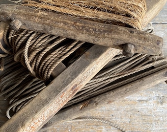 Pode incluir: Um enrolador de corda de madeira antigo com corda enrolada e um feixe de corda grossa de fibra natural. O enrolador é feito de madeira desgastada, mostrando sinais de idade e uso. A corda é de cor castanho claro.
