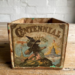 May include: A vintage wooden box with a faded label that reads "Centennial". The label features an illustration of a bald eagle flying over a cityscape with the Capitol building in the background.