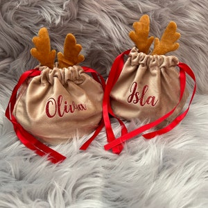 May include: Two gold-colored drawstring bags with red ribbon ties and brown reindeer antlers. The bags have the names "Olivia" and "Isla" embroidered in red glitter. The bags are on a fluffy, gray surface.